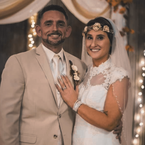 A smiling bride in a lace gown and flower crown standing with her groom in a tan suit at their indoor country wedding reception.