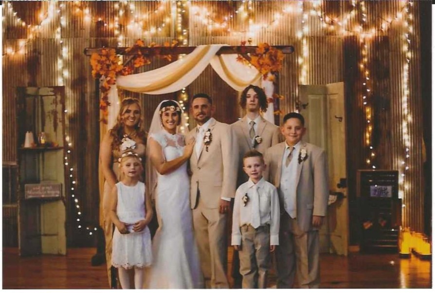 A bride, groom, and their wedding party including children posing together in front of a rustic wooden backdrop with warm fairy lights and white draping.