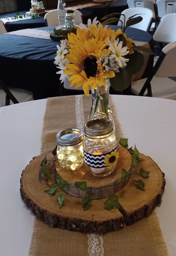 Rustic wedding centerpiece with sunflowers, daisies, and a fairy light mason jar on a wood slice