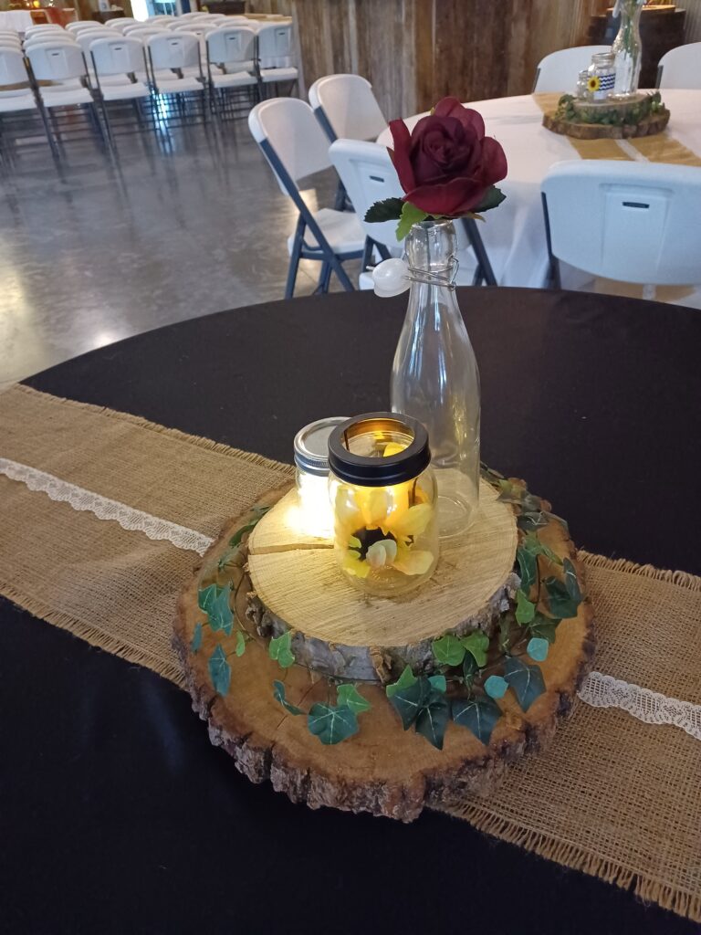 A rustic wedding centerpiece on a black tablecloth featuring a natural wood slice wrapped in ivy, a glowing mason jar, and a burgundy rose in a glass vase.