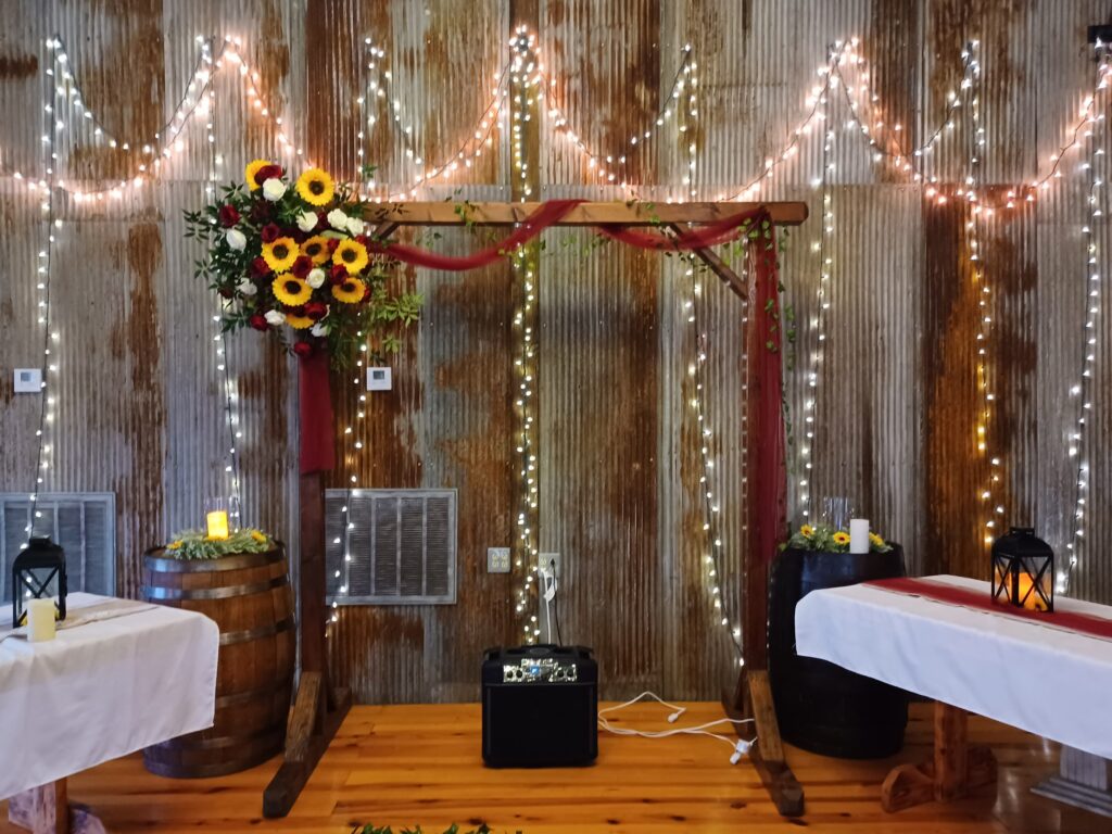 Wooden wedding arch with sunflowers and burgundy roses against a corrugated tin backdrop with fairy lights