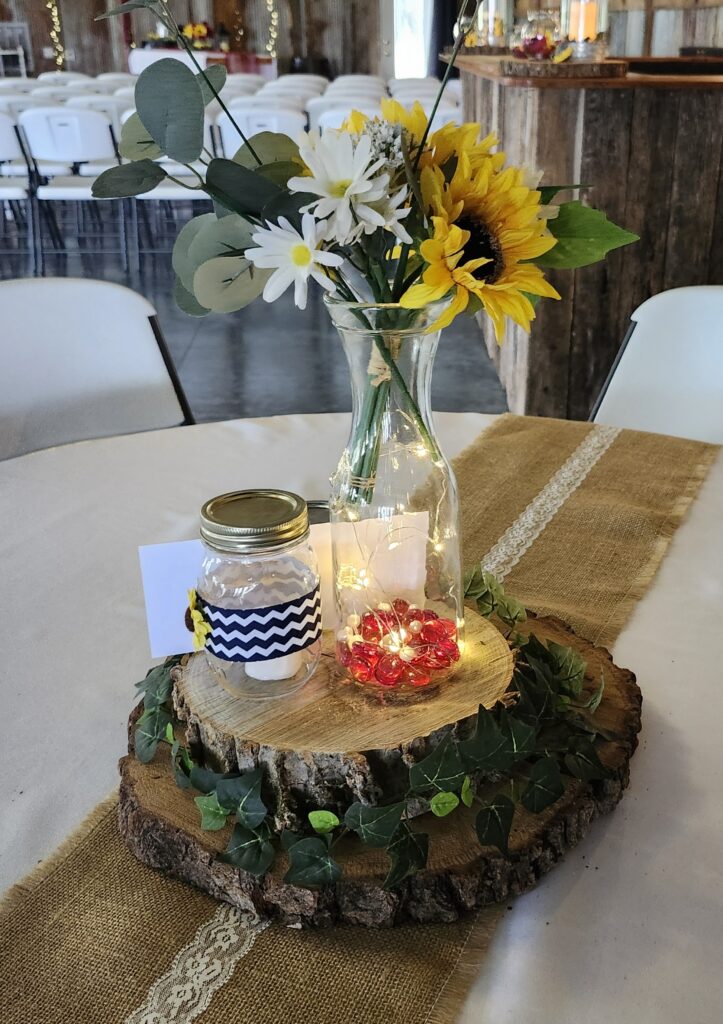 Rustic wedding centerpiece with sunflowers, white daisies, and a fairy light mason jar on a wood slice