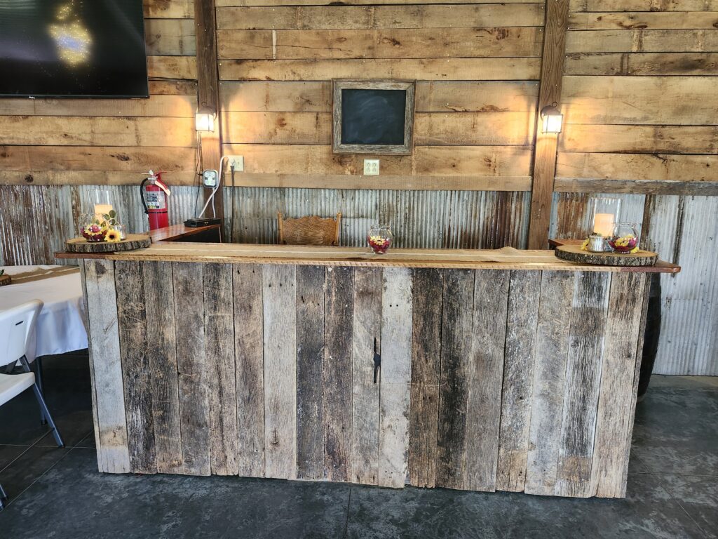 A rustic indoor wedding bar made of weathered wood planks decorated with sunflowers and candles against a corrugated metal wall.