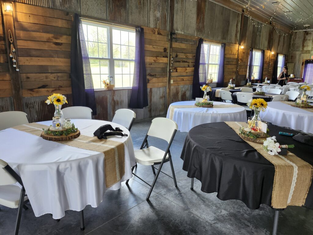 Indoor wedding reception setup with rustic wood walls, round tables, burlap runners, and sunflower centerpieces