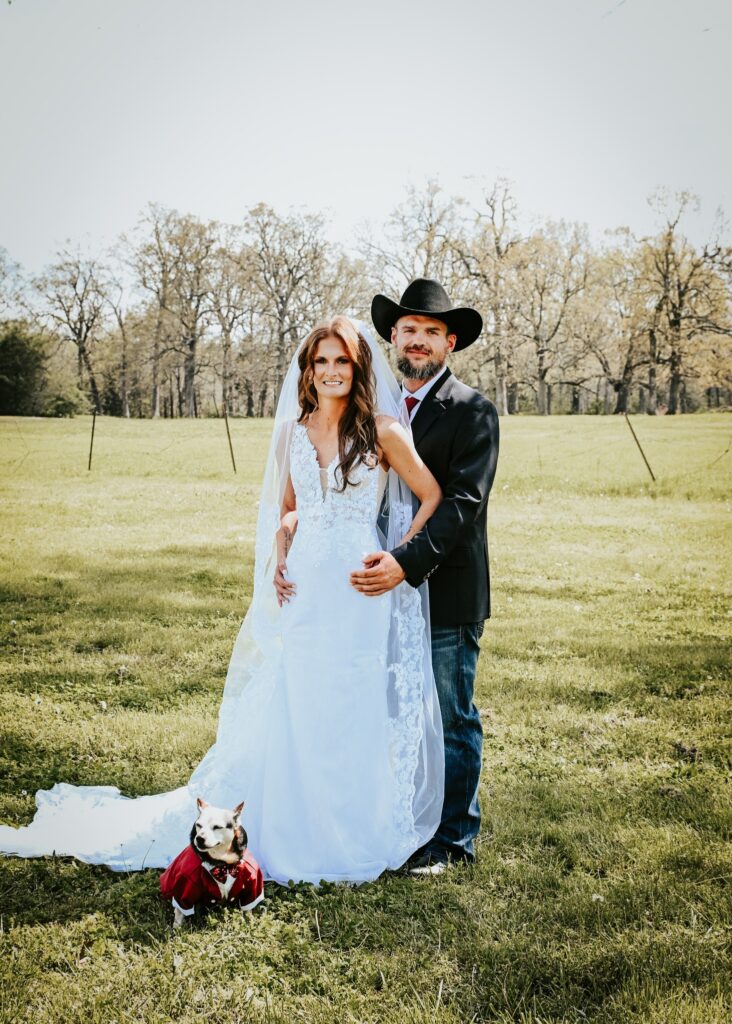 Bride and groom posing in the open field with a small dog wearing a wedding outfit for unique rustic wedding inspiration at Country Charm Venue.  Photo by Lash & Lens Studio, LLC.