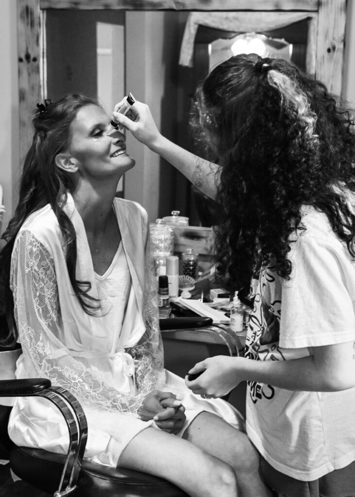 A black and white photo of a smiling bride sitting in a chair getting her hair and makeup done inside the Farmhouse bridal suite.