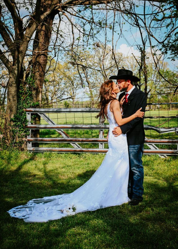 A bride in a lace wedding gown and a groom in a black cowboy hat kiss next to a metal farm gate on a grassy lawn.