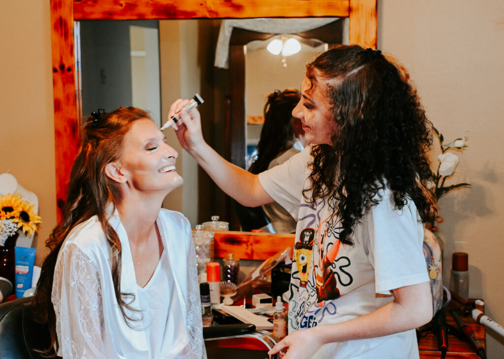 A bride in a white lace robe smiles while a makeup artist applies her makeup inside the Farmhouse bridal suite.