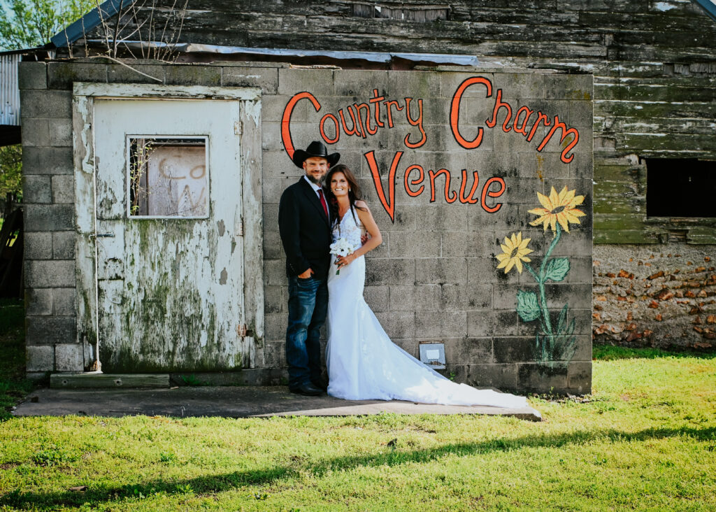 A bride in a lace wedding gown and a groom in a black cowboy hat pose in front of the rustic wooden barn with the hand-painted Country Charm Venue sign and sunflowers.
