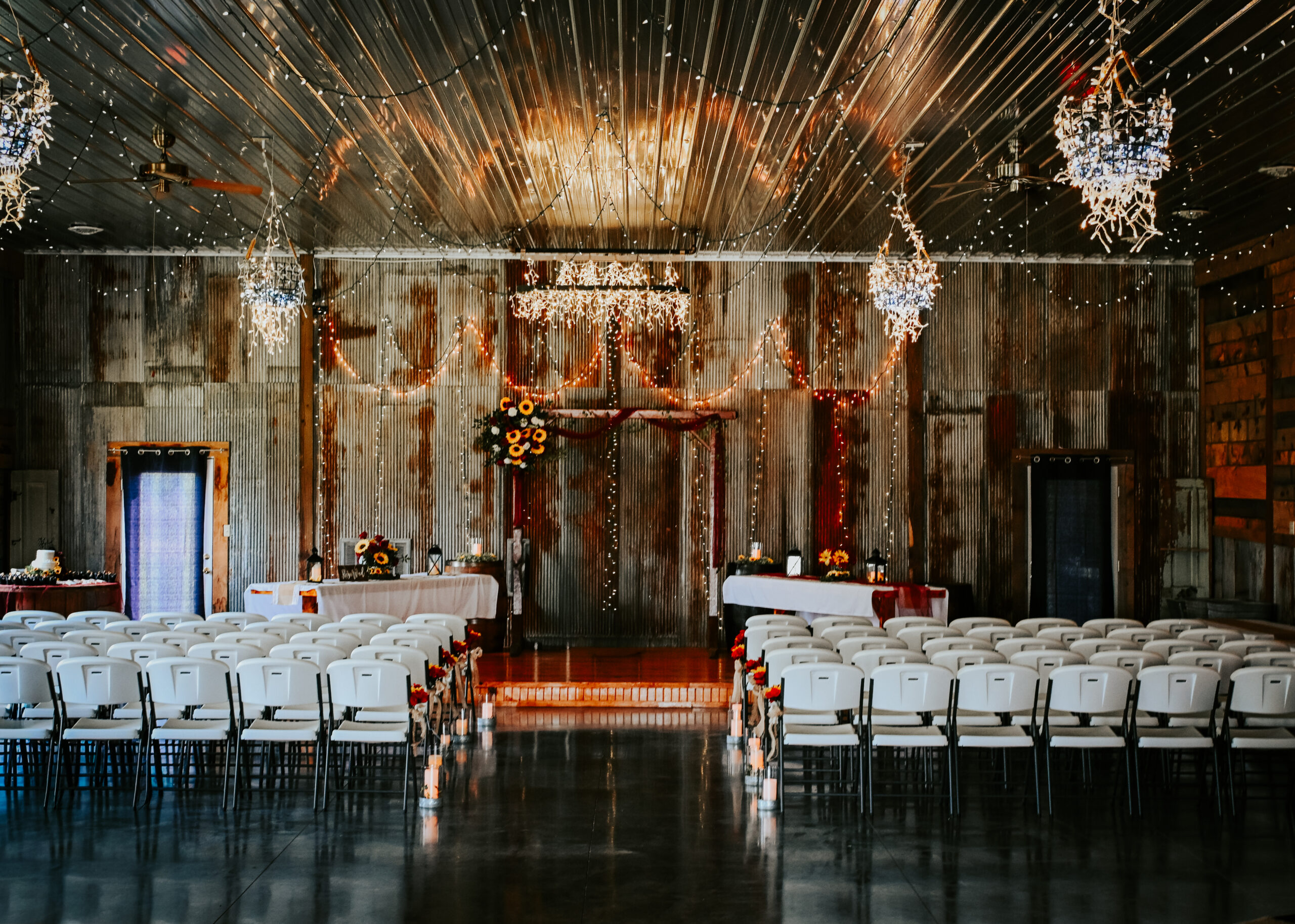 n empty rustic indoor wedding ceremony venue hall with rows of white chairs, a central aisle, glowing chandeliers, and a wooden altar backdrop.