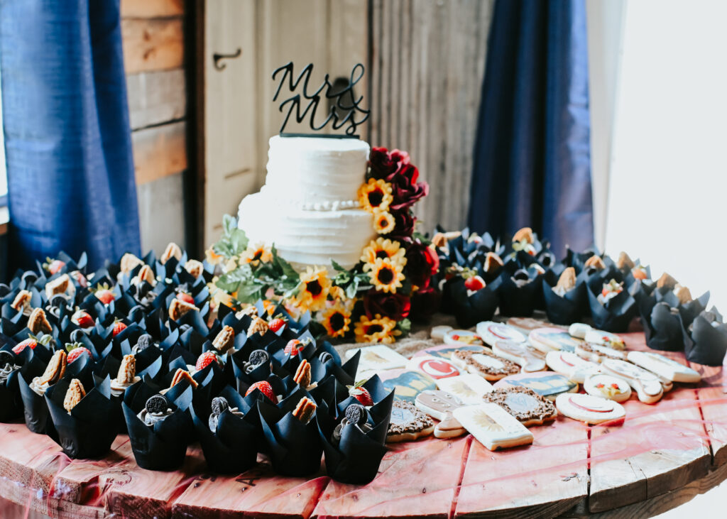 A three-tiered white wedding cake with a Mr. and Mrs. topper sits next to cupcakes and custom sugar cookies on a rustic wooden dessert table.