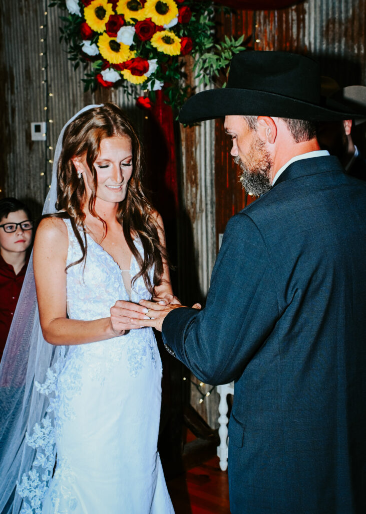 A groom wearing a black cowboy hat places a ring on the bride's finger during a rustic indoor wedding ceremony.