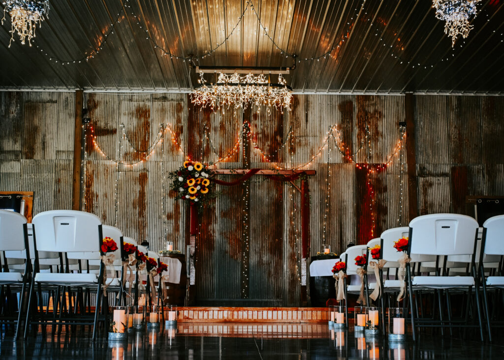 An indoor rustic wedding ceremony setup with string lights, chandeliers, a wooden altar arch, and rows of white chairs lined with flower petals.