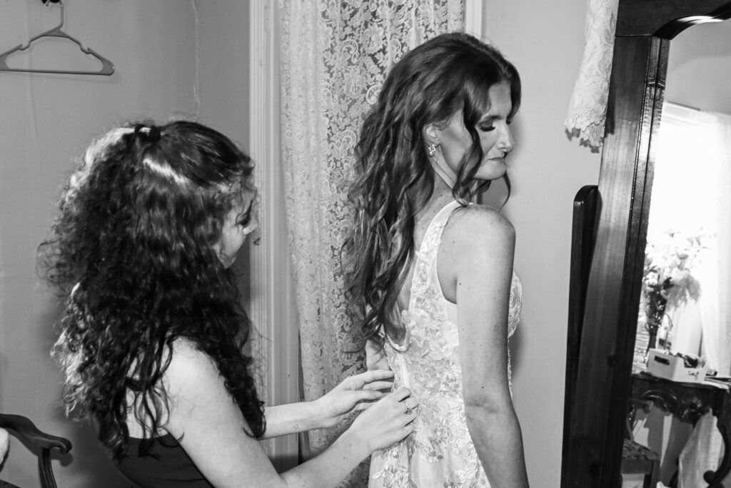 A black and white photo of a bride having the back of her lace wedding gown buttoned up by a bridesmaid inside the Farmhouse bridal suite.
