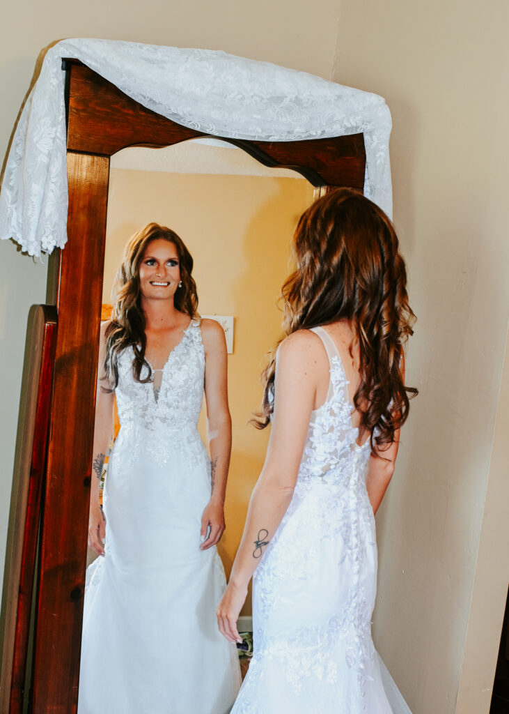 A bride in a lace wedding dress smiles at her reflection in a full-length wooden mirror inside the Farmhouse bridal suite.