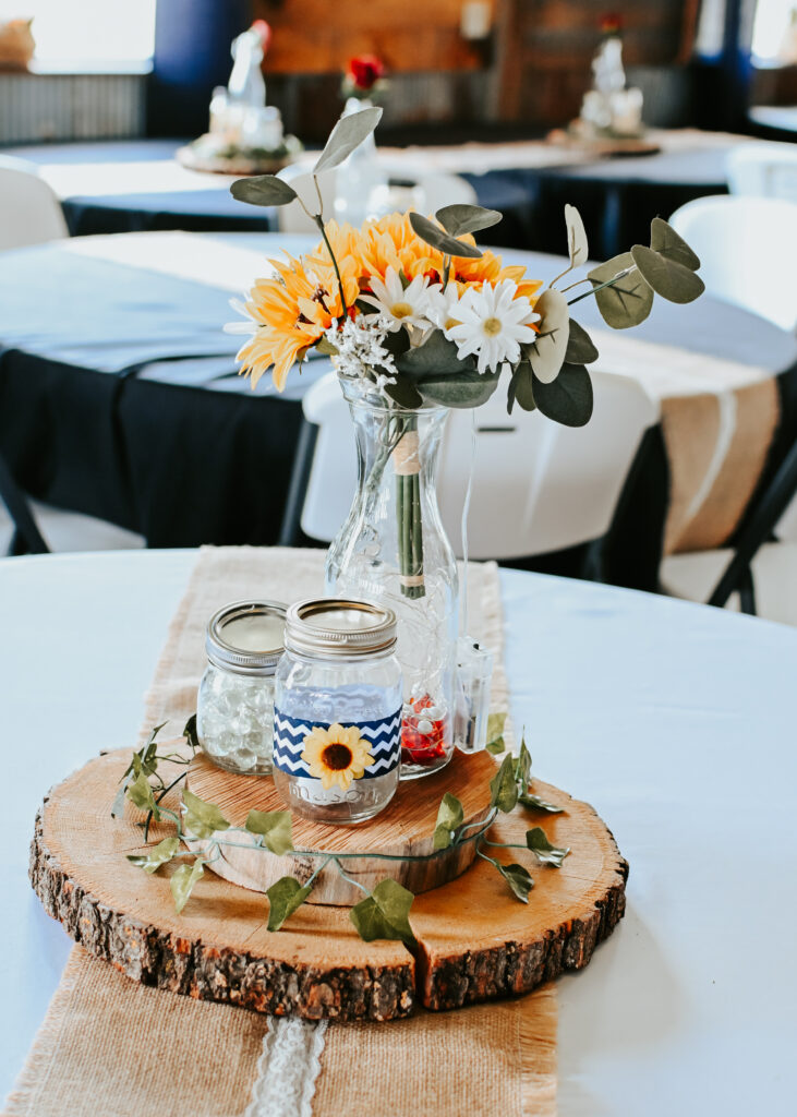 A wedding reception table centerpiece with a sunflower and daisy floral arrangement in a glass vase sitting on a rustic wood slice with mason jars.