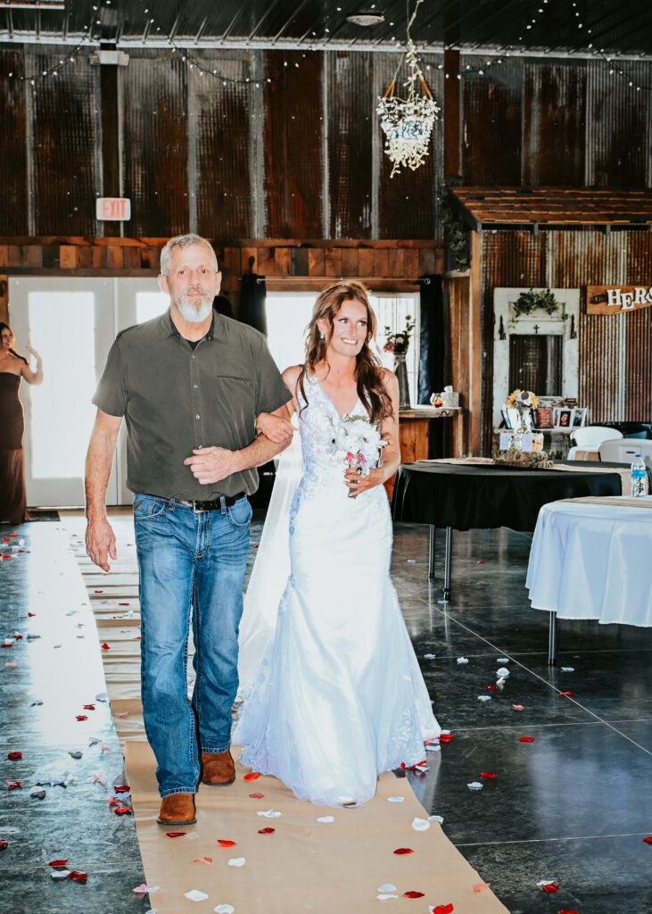 A bride in a lace wedding gown is walked down the aisle by a man during an indoor rustic wedding ceremony with red flower petals on the floor.