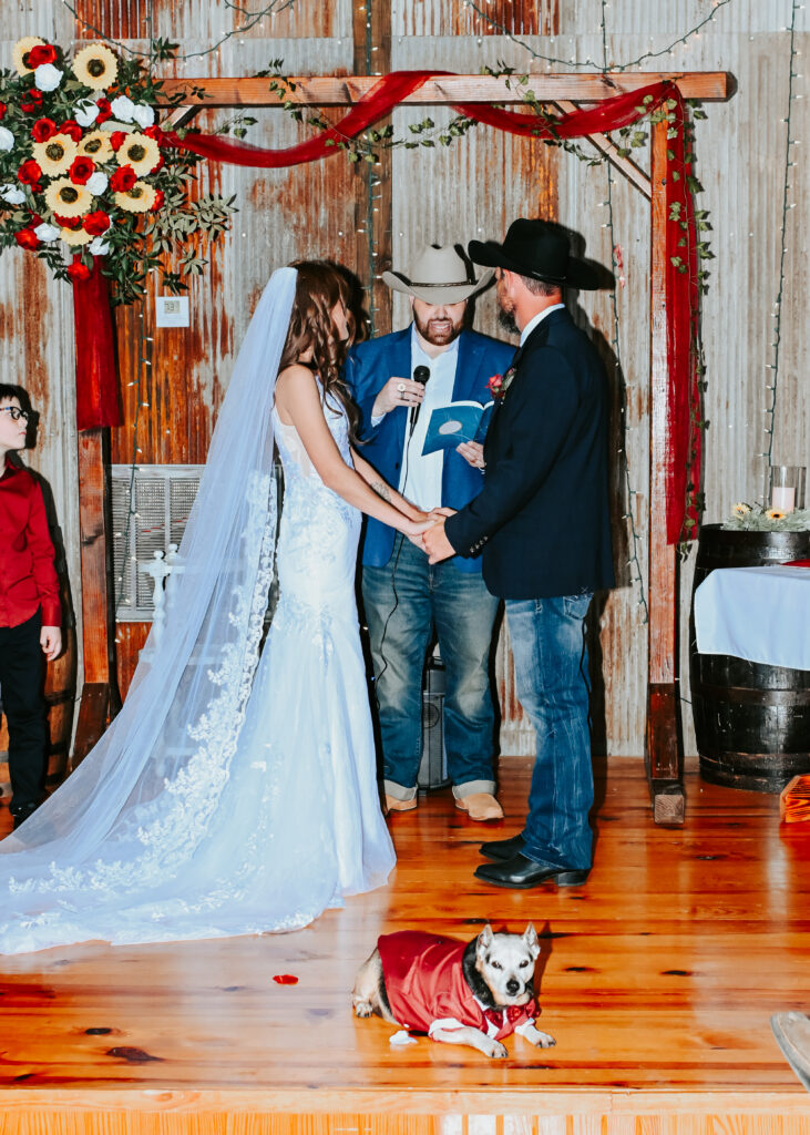 A bride and groom hold hands at the altar during an indoor wedding ceremony with an officiant and a small dog wearing a red outfit sitting on the floor.