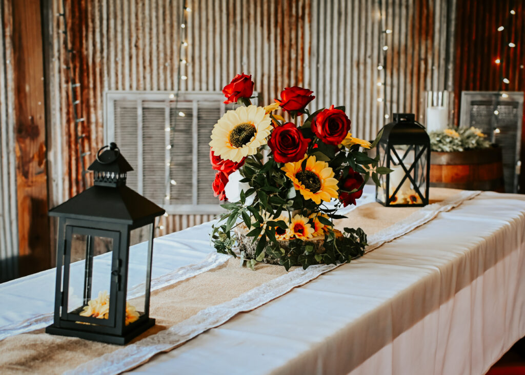 A wedding reception table setup with a white tablecloth, burlap and lace runner, a centerpiece of sunflowers and red roses, and black lanterns.
