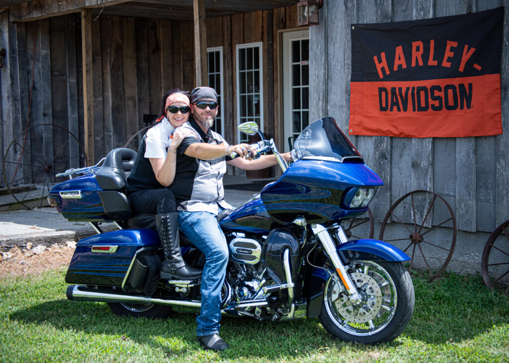A couple sits on their Harley Davidson in front of the porch of Country Charm Venue with a Harley Davidson flag behind them.